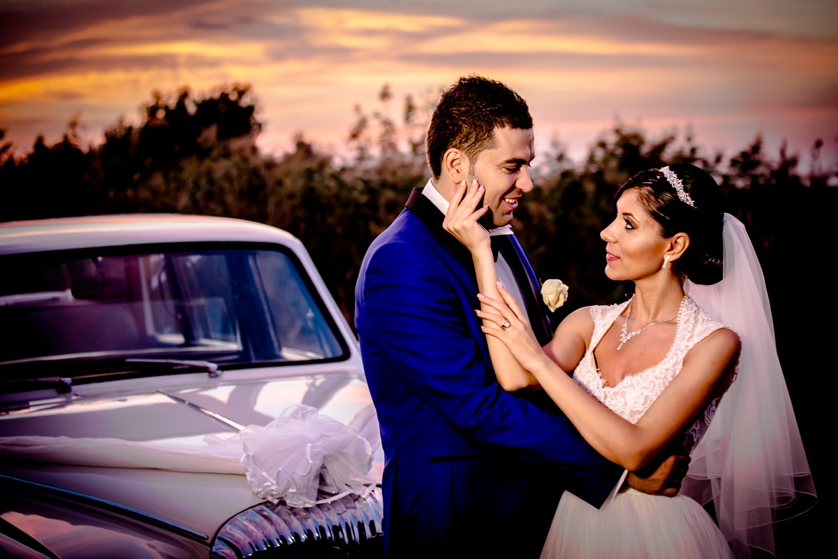Mire și mireasă fotografiați la apus, lângă o mașină de epocă, într-un moment elegant și romantic